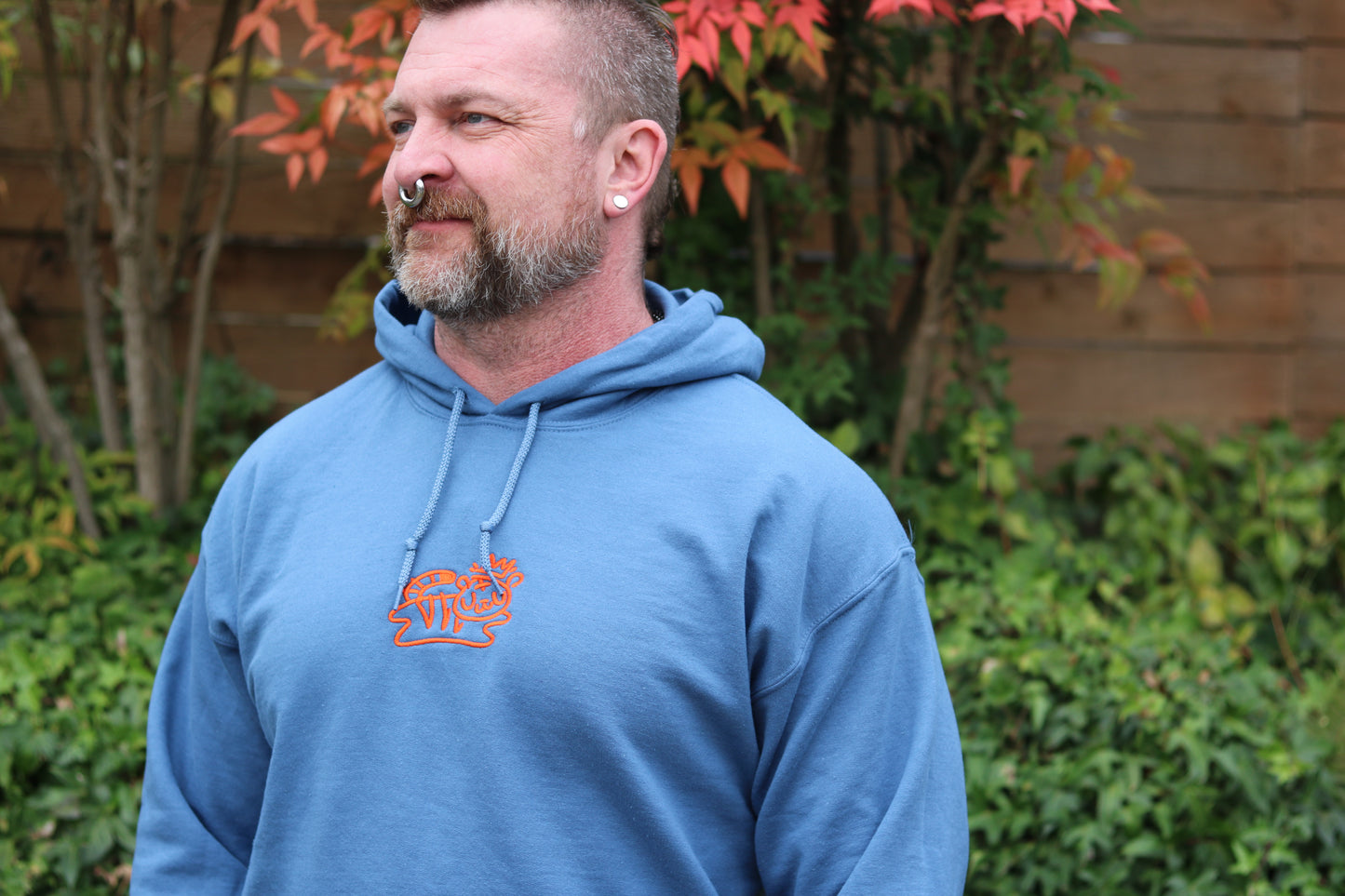 handsome man with nose ring and beard wearing royal blue pullover hoodie with bolt-wear tiger logo standing in front of leaves and ivy, fence in background