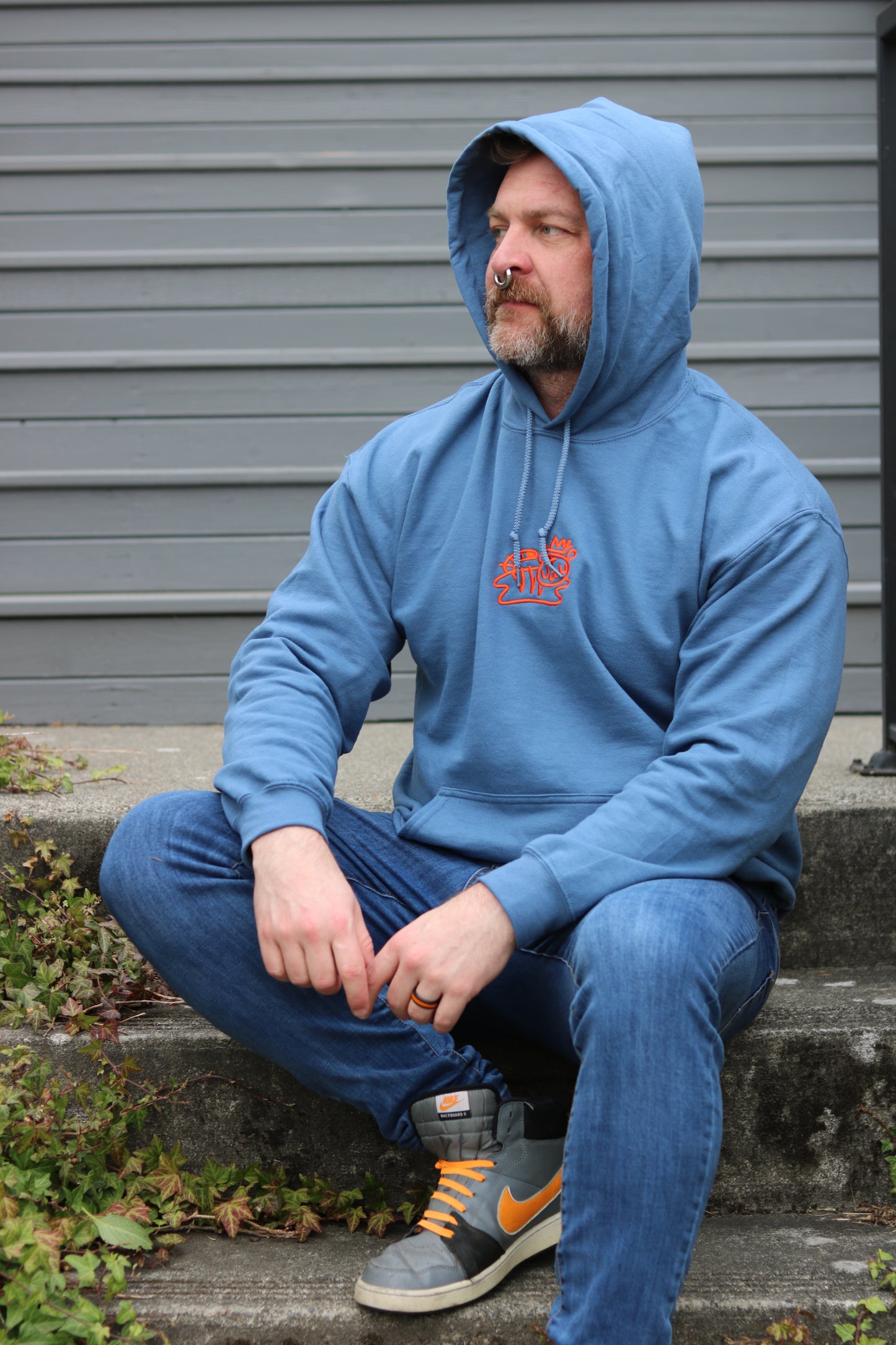 handsome man with nose ring and beard wearing royal blue pullover hoodie with bolt-wear tiger logo sitting on stair with overgrown ivy
