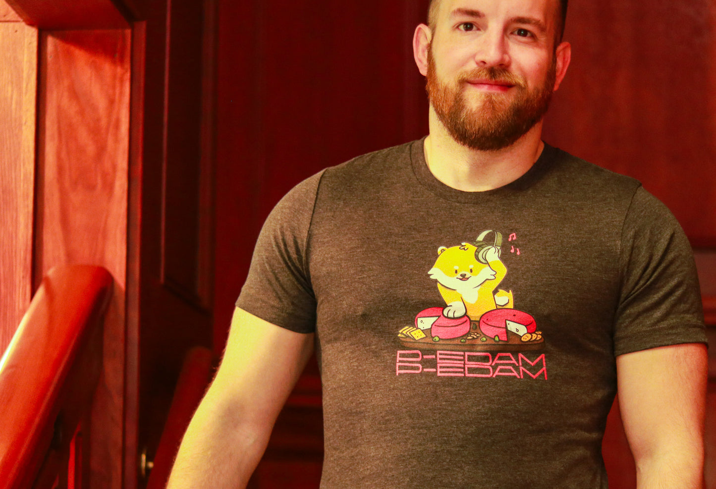 Bearded person walking down stairs wearing dark grey heather P-Edam t-shirt featuring illustration of Pomeranian playing two wheels of edam cheese like DJ tables on a charcuterie