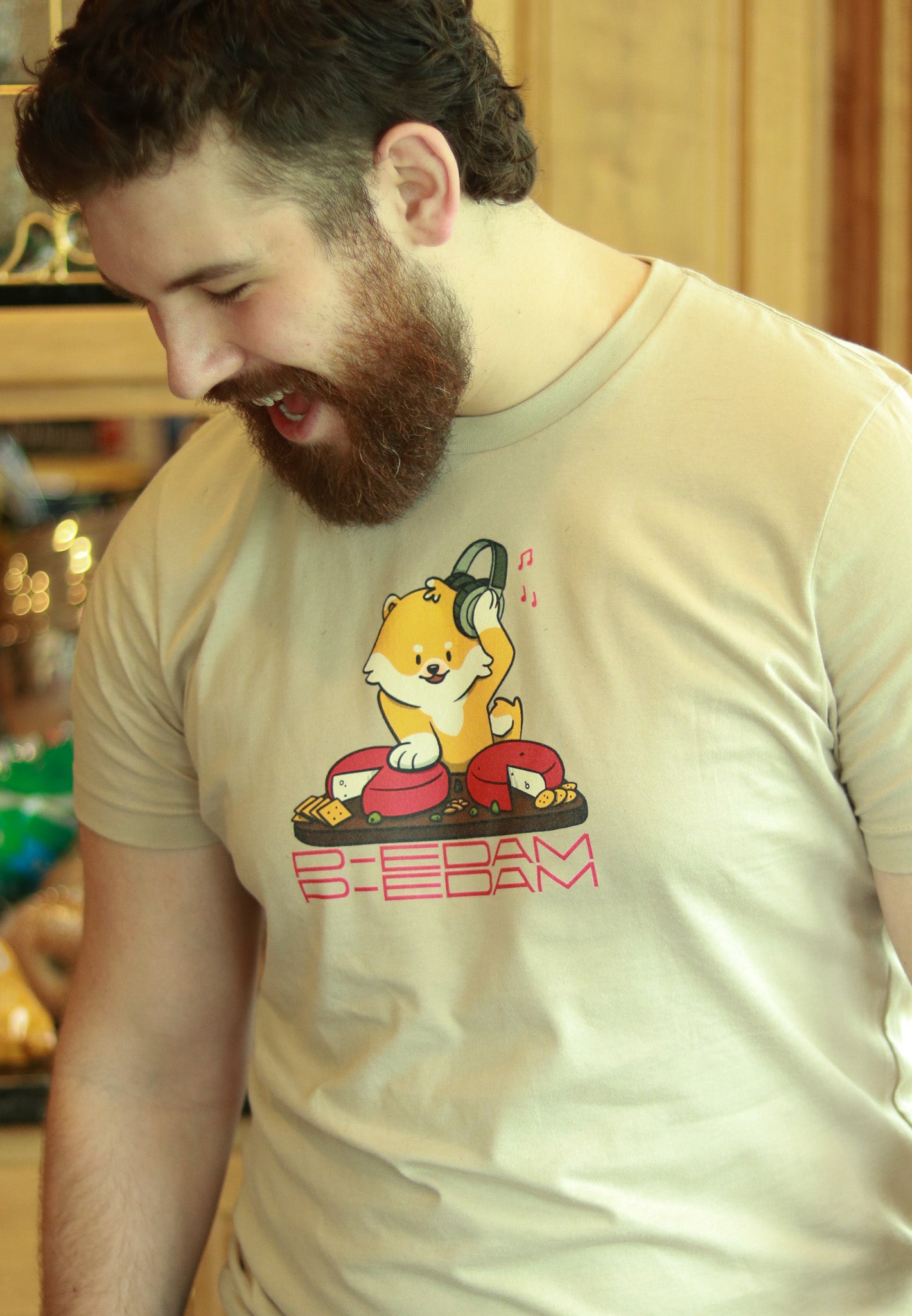 bearded person in kitchen wear tan P-Edam t-shirt featuring illustration of Pomeranian playing two wheels of edam cheese like DJ tables on a charcuterie