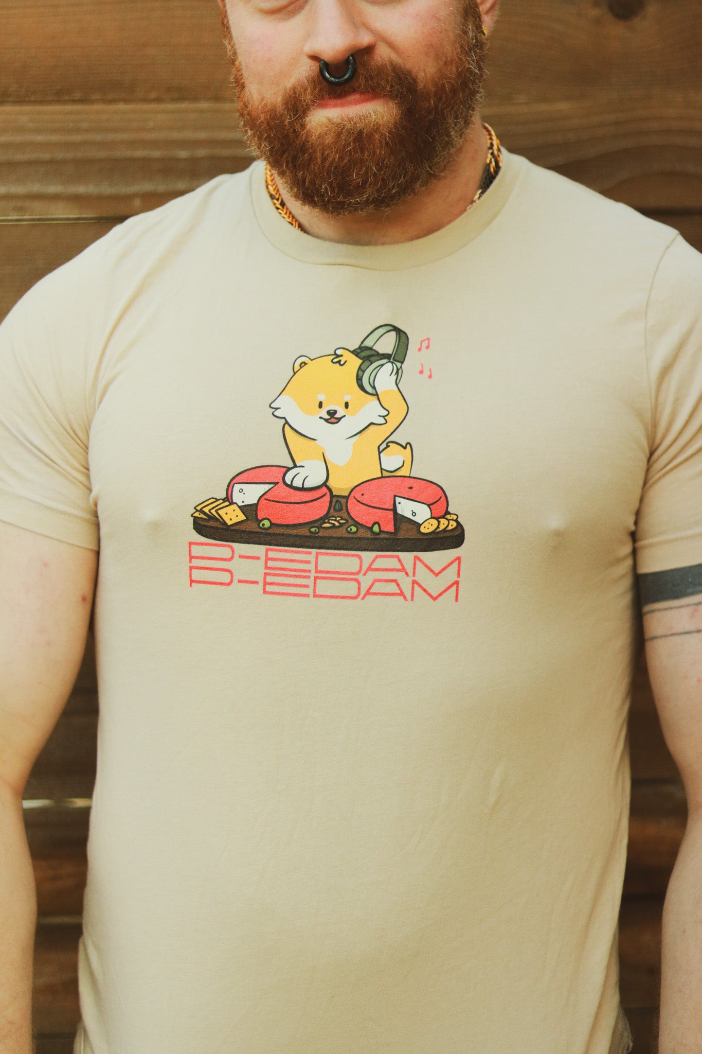 Ginger with beard standing in front of wooden wall wearing tan P-Edam t-shirt featuring illustration of Pomeranian playing two wheels of edam cheese like DJ tables on a charcuterie