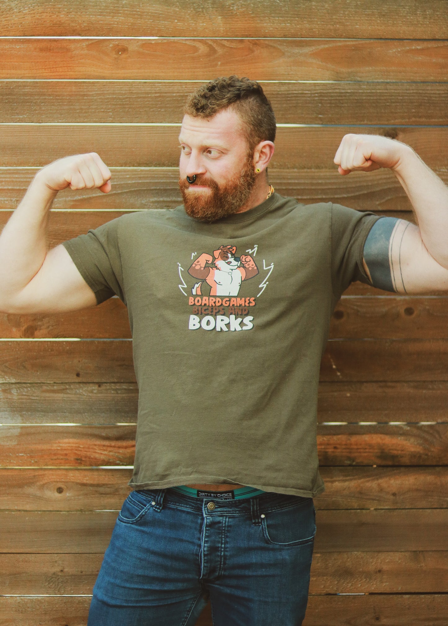 Ginger with beard mohawk and piercings flexing in front of a wall wearing an olive green shirt with orange and white dog, text underneath reading 'Boardgames Biceps and Borks"