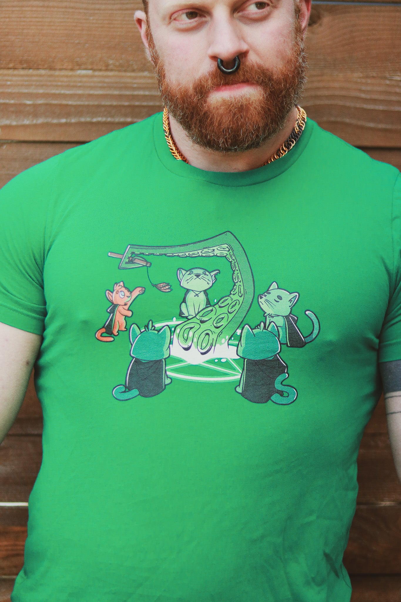 Ginger with beard wearing green shirt featuring 5 cats summoning a tentacle, one an orange cat playing with a feather toy held by the tentacle