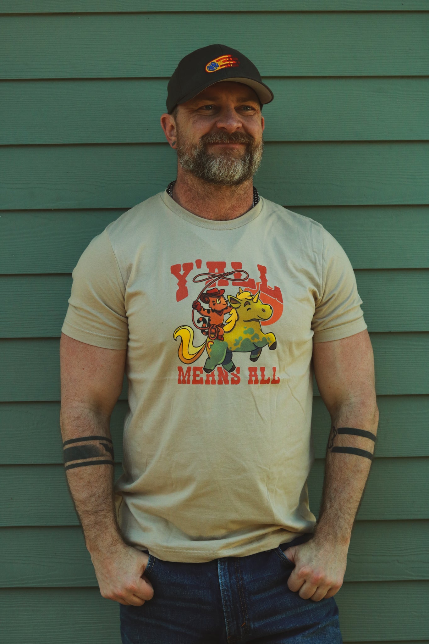 light tan tshirt worn by bearded guy with d20 comet hat from bolt-wear in front of green wall, displaying tiger riding unicorn in rainbow colors and the text "y'll means all"