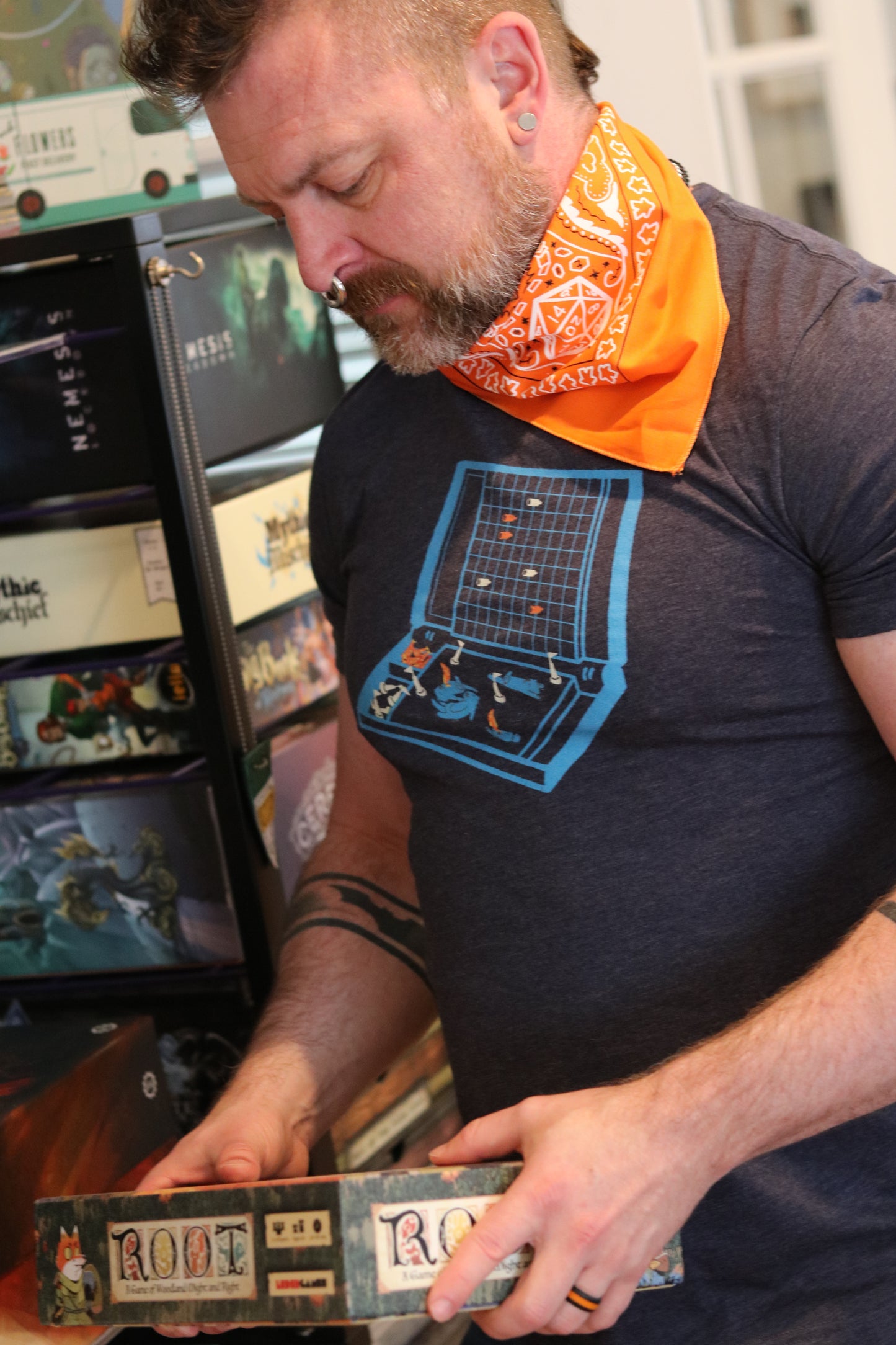 Handsome bearded man with nose ring holding root board game, wearing orange D20 dungeons and dragons theme bandana around neck
