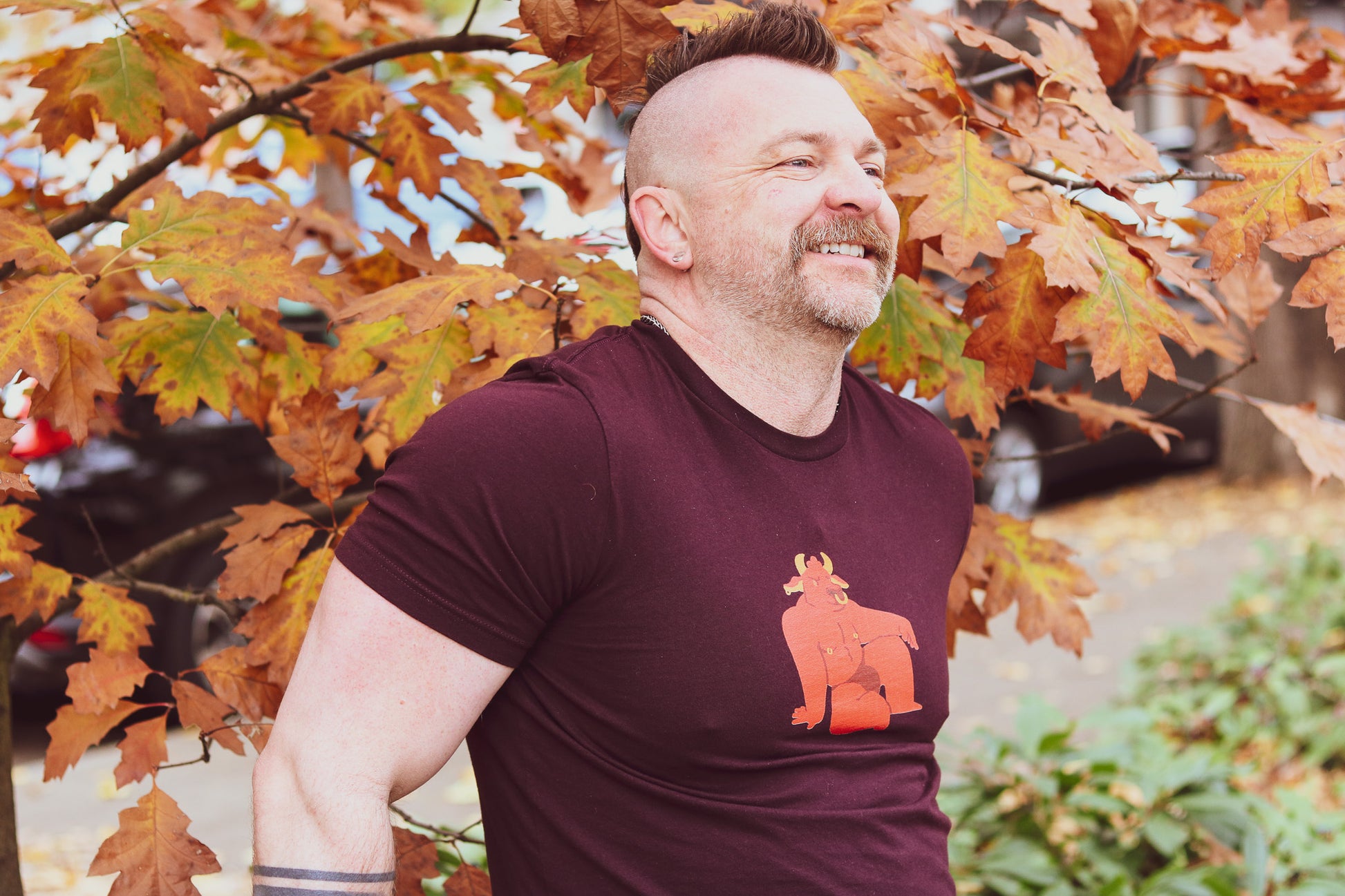 handsome man with mohawk and beard wearing oxblood black tshirt featuring sexy red bull furry with nose and nipple rings in a jock strap, sitting in open looking pose, standing in front of leaves with bicep flexed.