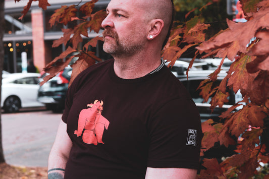 handsome man with mohawk, pierced ears, and beard wearing oxblood black tshirt featuring sexy red bull furry with nose and nipple rings in a jock strap, sitting in open looking pose, standing in front of leaves with bolt-wear tiger logo showing on sleeve.