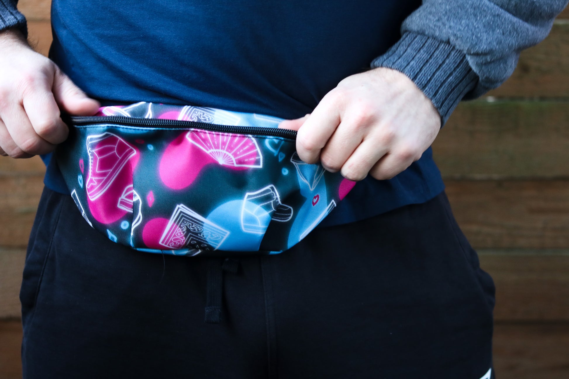 Front view of pink and blue fanny pack featuring images of high top shoes fans and bandanas, model wearing sweatshirt holding the corners