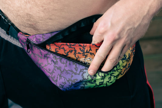 All over print fanny pack featuring rainbow bear pattern in style of Keith Haring with shirtless model opening interior pocket