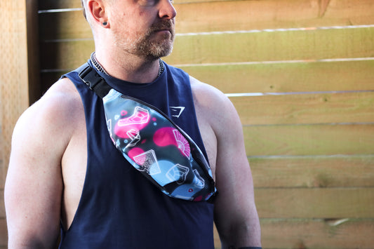 Front view of pink and blue fanny pack featuring images of high top shoes fans and bandanas, model with beard and earing wearing bag over the shoulder wearing sleeveless shirt
