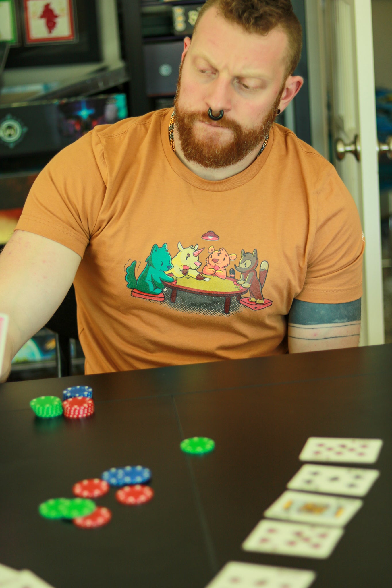 Dusty Orange t-shirt featuring 4 animals, a wolf, a unicorn, a tiger, and a racoon, playing cards around a table, worn by bearded ginger with mohawk and tattoos playing cards.