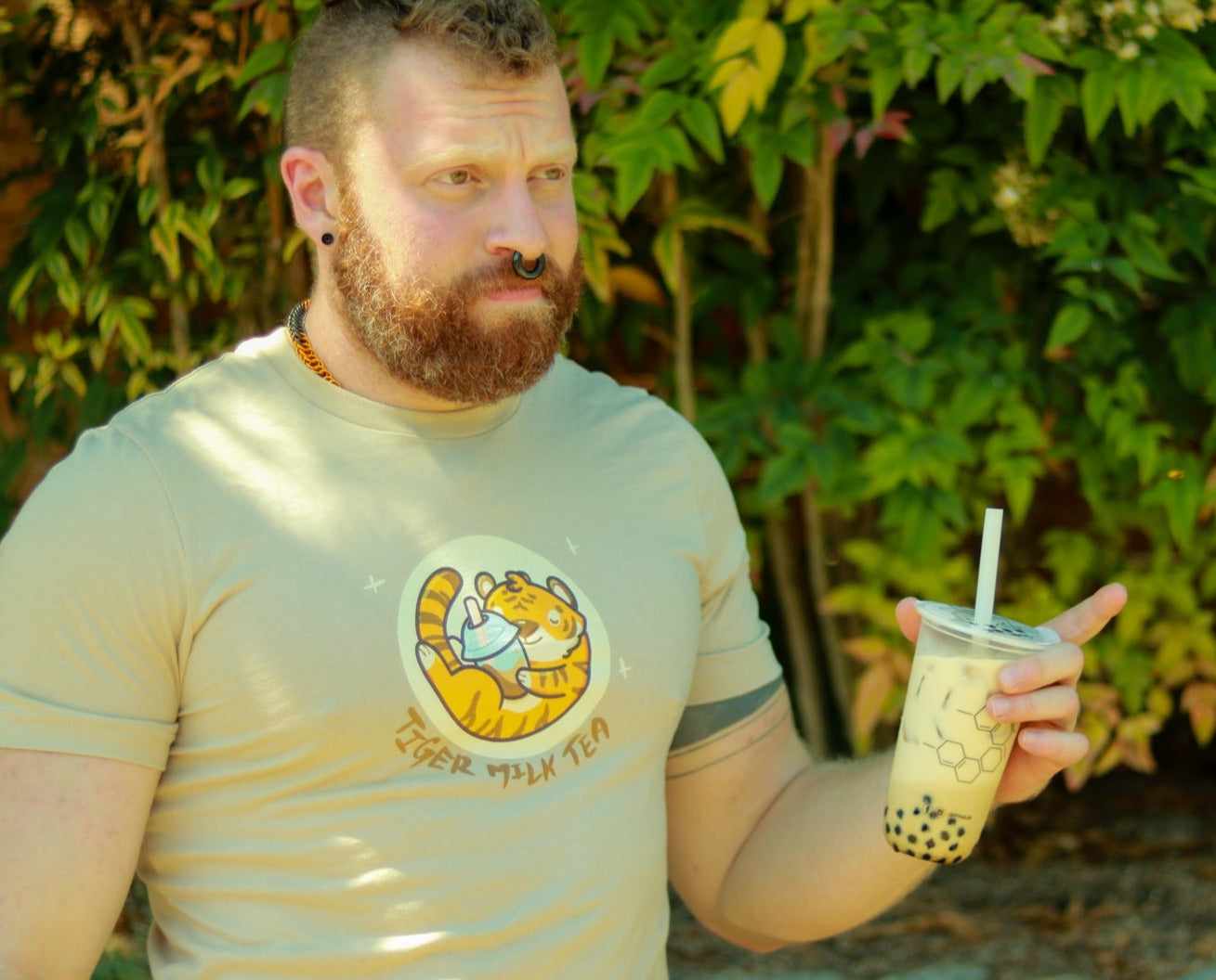Muscular bearded ginger with earrings and nose holding bubble tea wearing tan t-shirt featuring image of tiger holding bubble tea, with the words tiger milk tea underneath.