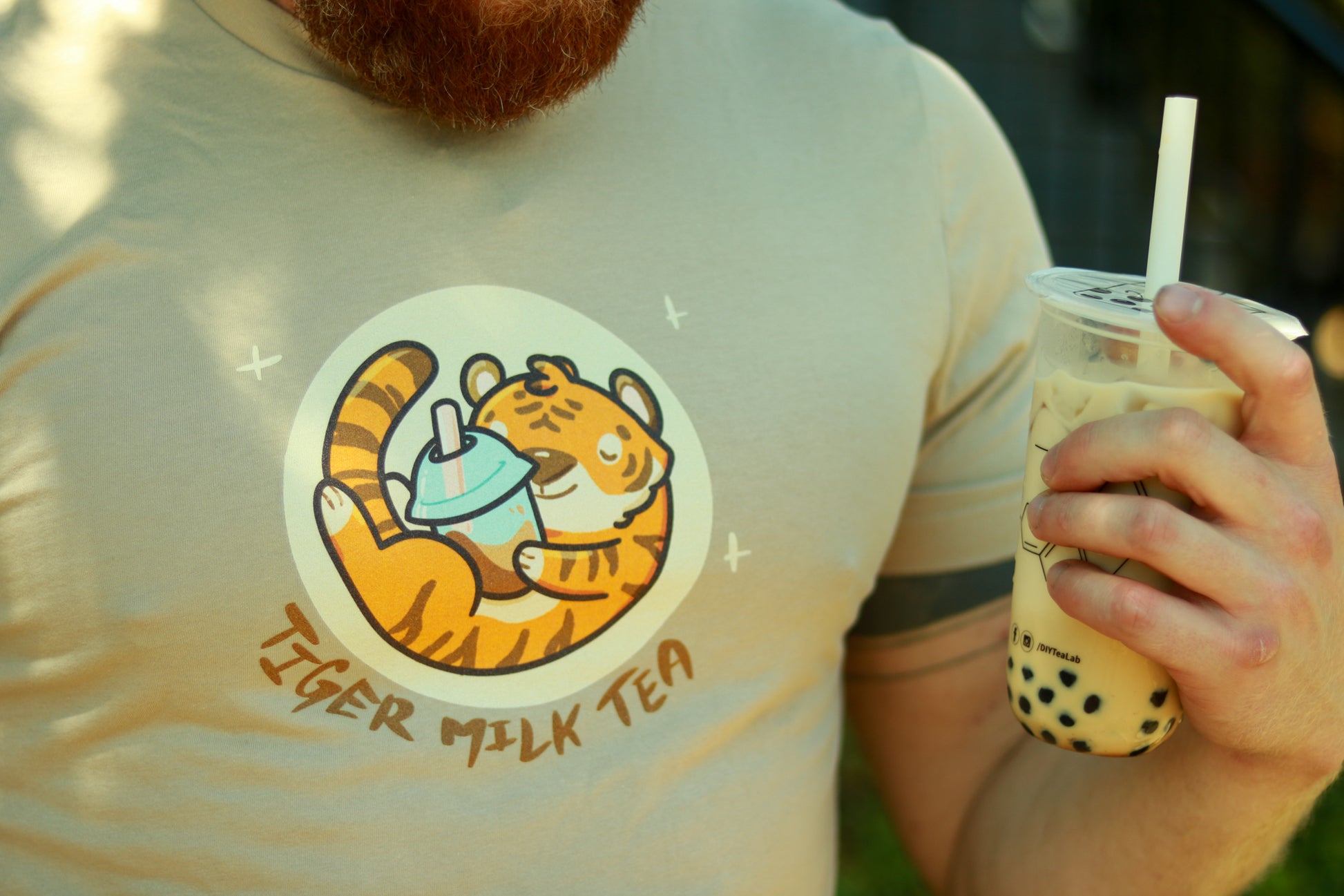 Close up of chest with hand holding bubble tea, person is wearing tan shirt featuring  image of tiger holding bubble tea, with the words tiger milk tea underneath.