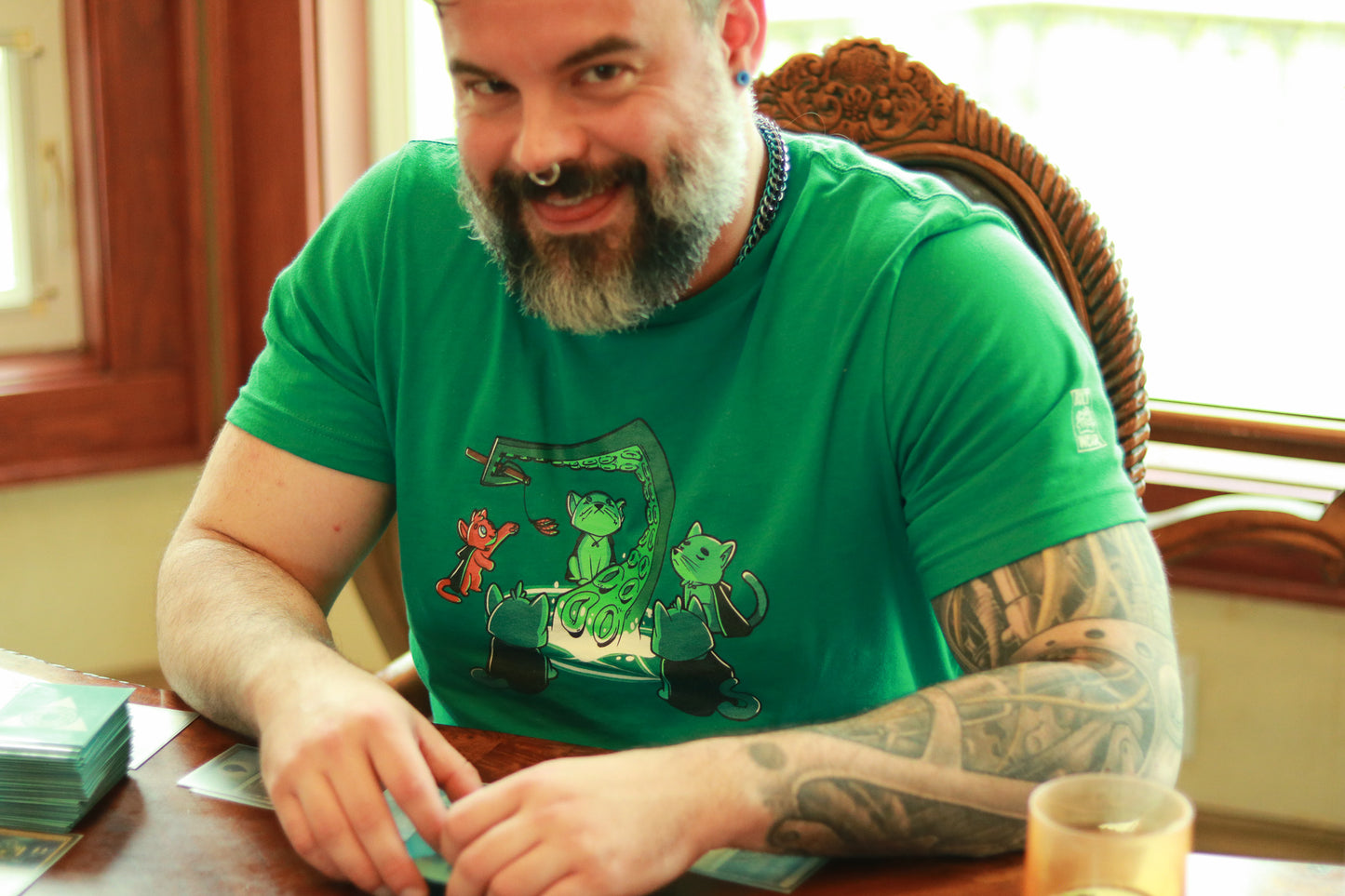 Devious man with beard playing magic the gathering wearing t-shirt featuring5 cats summoning a tentacle, one an orange cat playing with a feather toy held by the tentacle