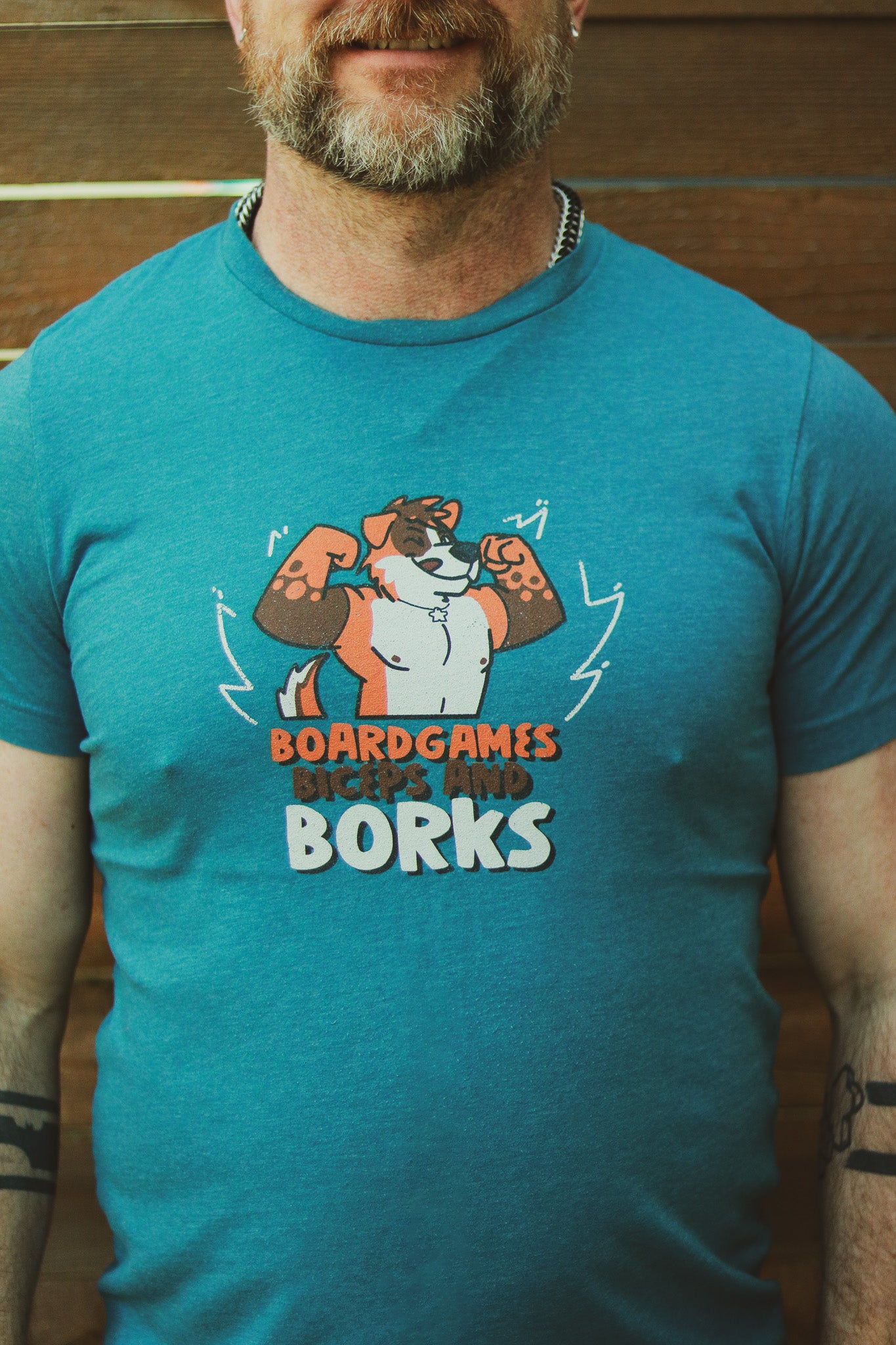 Man with a beard wearing blue heather shirt with orange and white flexing dog, text underneath reading 'Boardgames Biceps and Borks" 