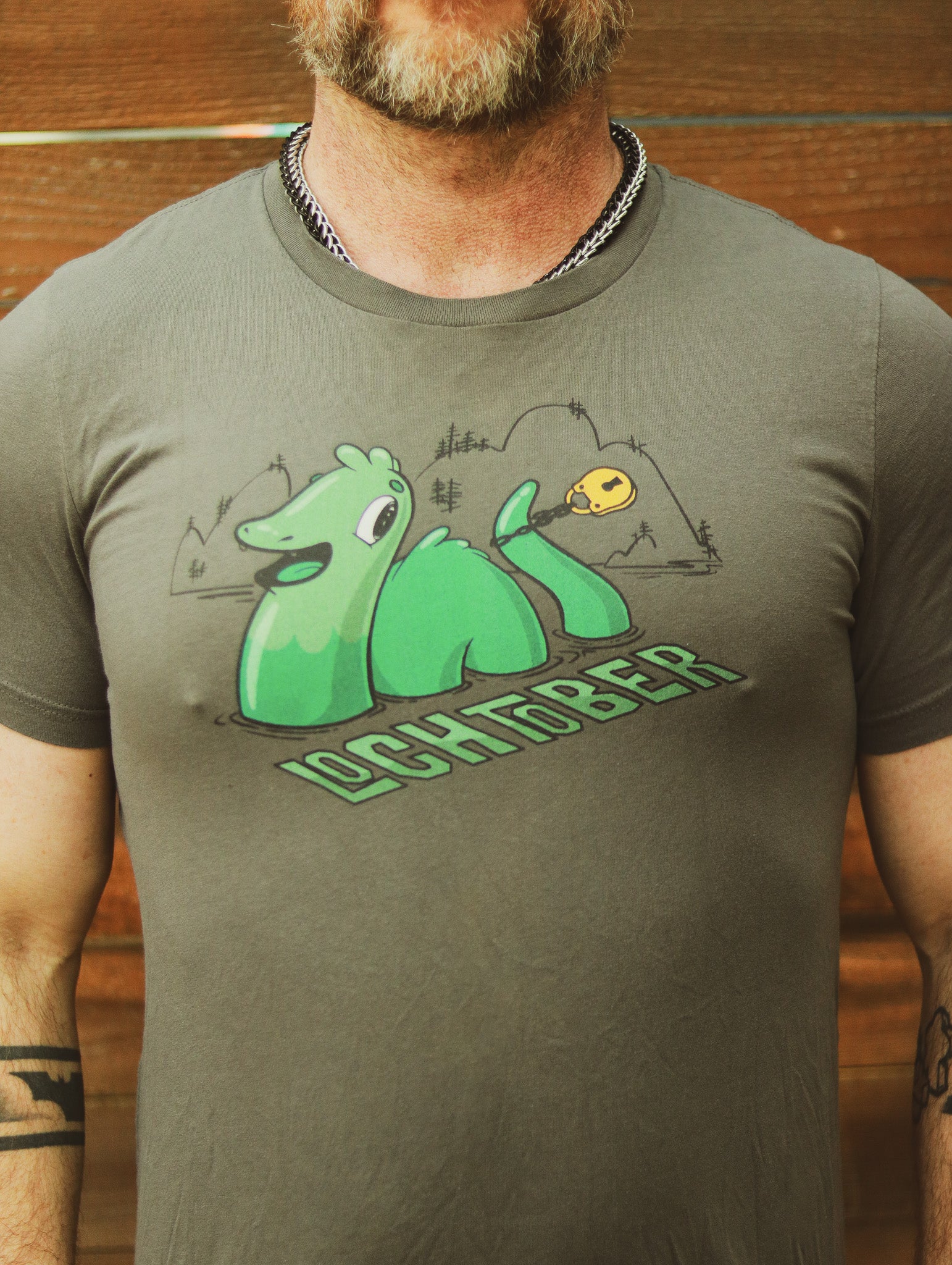 Grey Lochtober shirt featuring lochness monster with lock around tail, worn by bearded man standing in front of wooden wall..