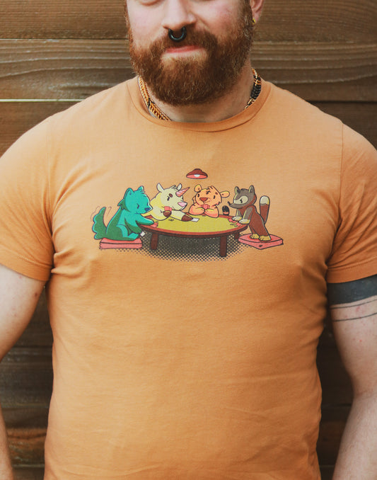 Dusty Orange t-shirt featuring 4 animals, a wolf, a unicorn, a tiger, and a racoon, playing cards around a table, worn by bearded ginger with tattoos standing in front of wooden wall..