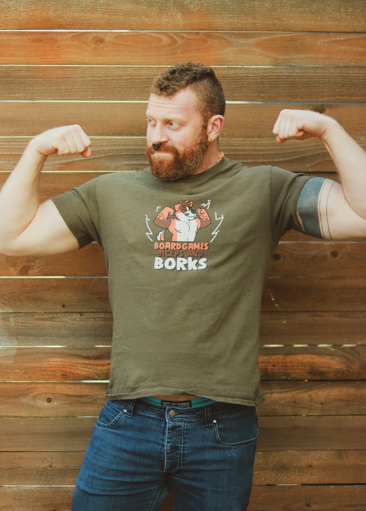 Ginger with beard mohawk and piercings flexing in front of a wall wearing an olive green shirt with orange and white dog, text underneath reading 'Boardgames Biceps and Borks" 