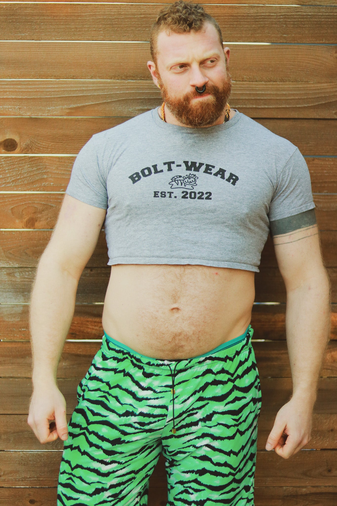 Ginger with beard and piercings standing in front of wooden wall wearing grey heather crop top reading Bolt-wear Est. 2022 featuring the boltwear logo and green tiger stripped pants