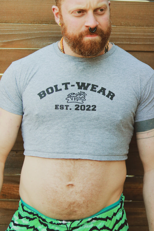 Ginger with beard and piercings standing in front of wooden wall wearing grey heather crop top reading Bolt-wear Est. 2022 featuring the boltwear logo
