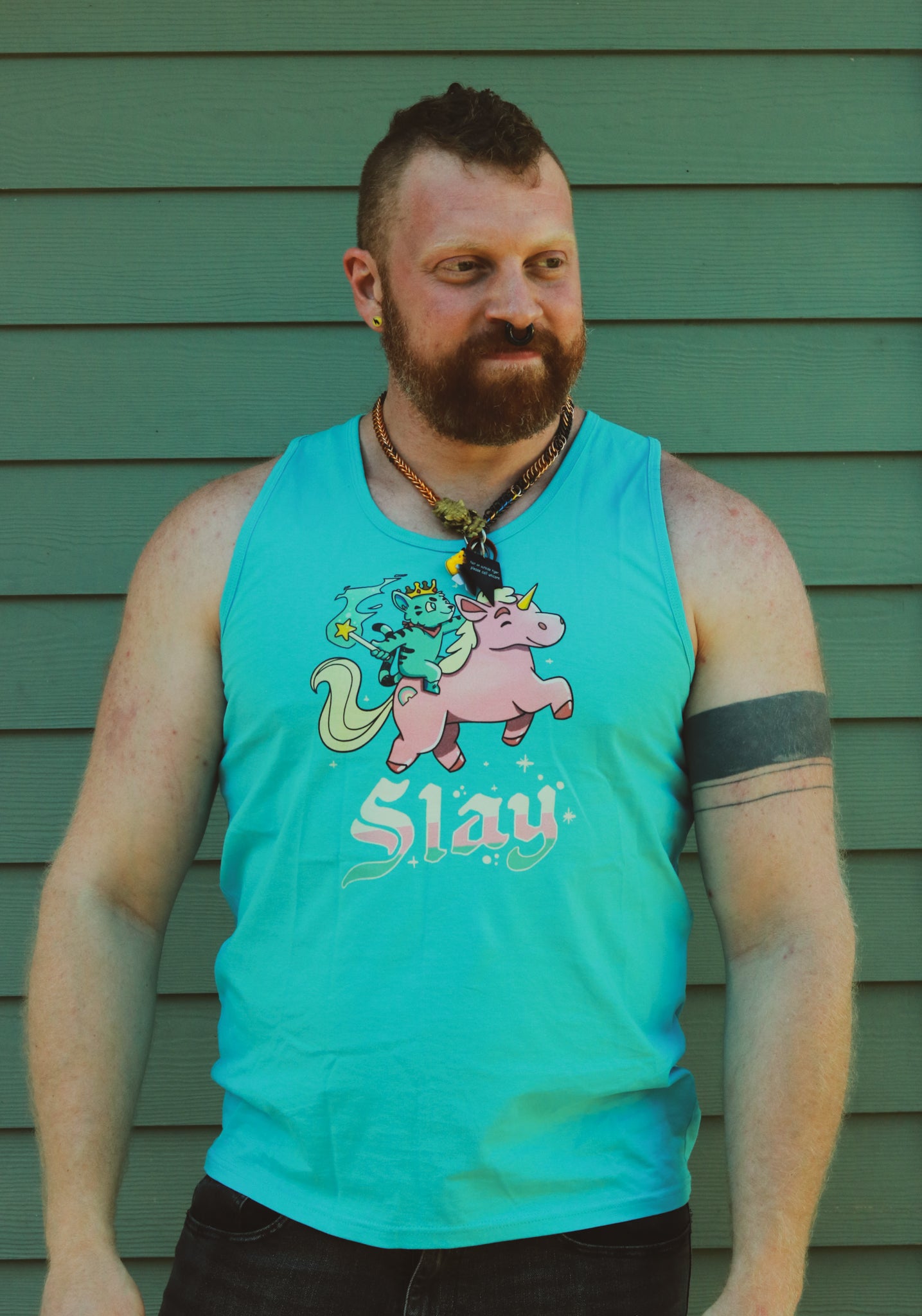 a light blue tank top with blue tiger riding pink unicorn, with the words slay below the image, worn by bearded hairy ginger with an arm band tattoo..