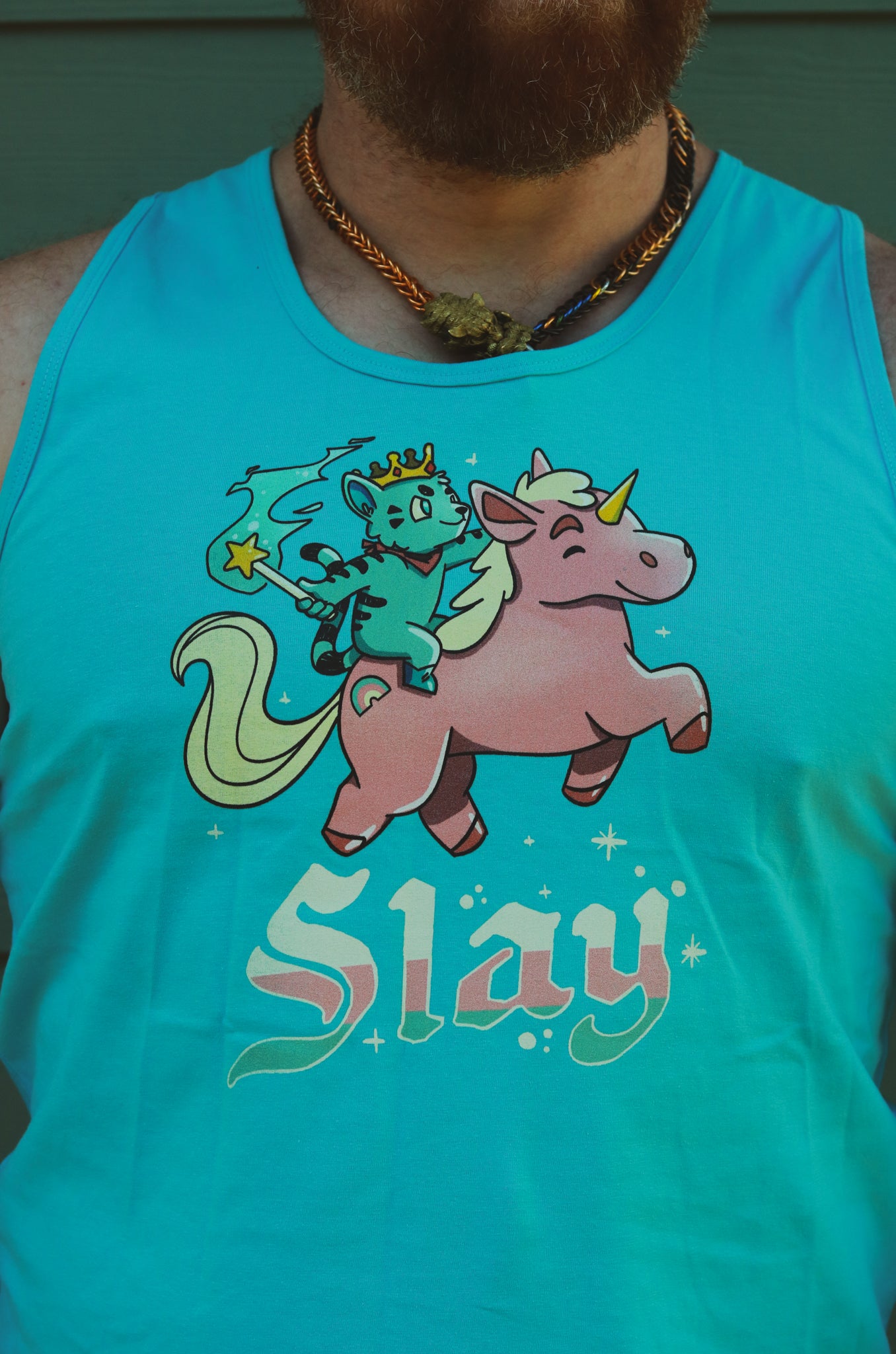 a light blue tank top with blue tiger riding pink unicorn, with the words slay below the image, worn by bearded hairy guy.