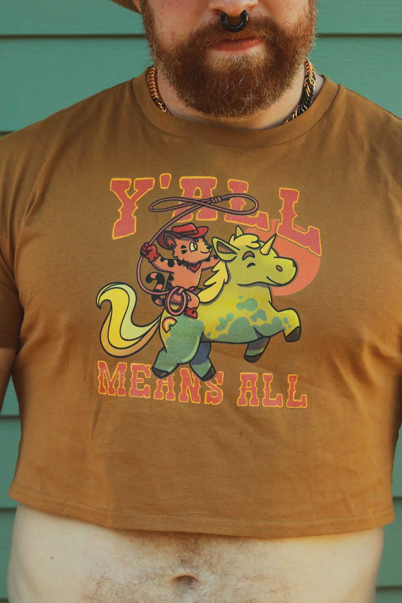 tan crop top worn by bearded ginger, displaying tiger riding unicorn in rainbow colors and the text "y'll means all"