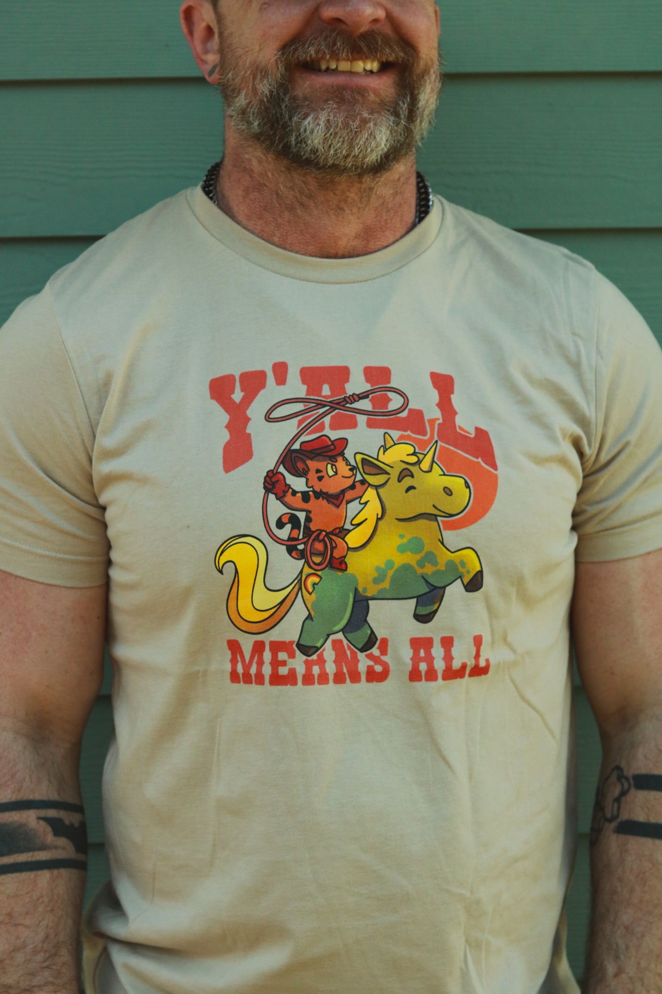 light tan tshirt top worn by bearded guy, displaying tiger riding unicorn in rainbow colors and the text "y'll means all"