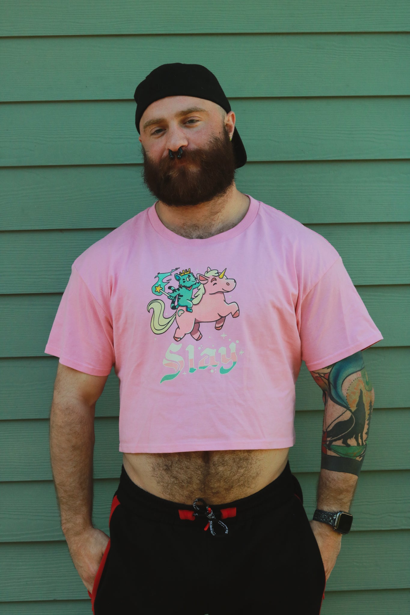 pink crop top with blue tiger riding pink unicorn, with the words slay below the image, worn by bearded hairy guy with backwards hat, in front of green wall..