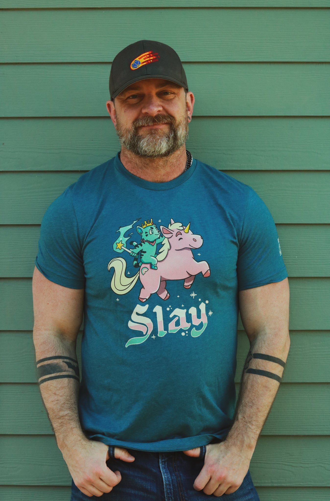 a blue heather tshirt with blue tiger riding pink unicorn, with the words slay below the image, worn by bearded hairy guy wearing a comet d20 hat in front of a green wall..