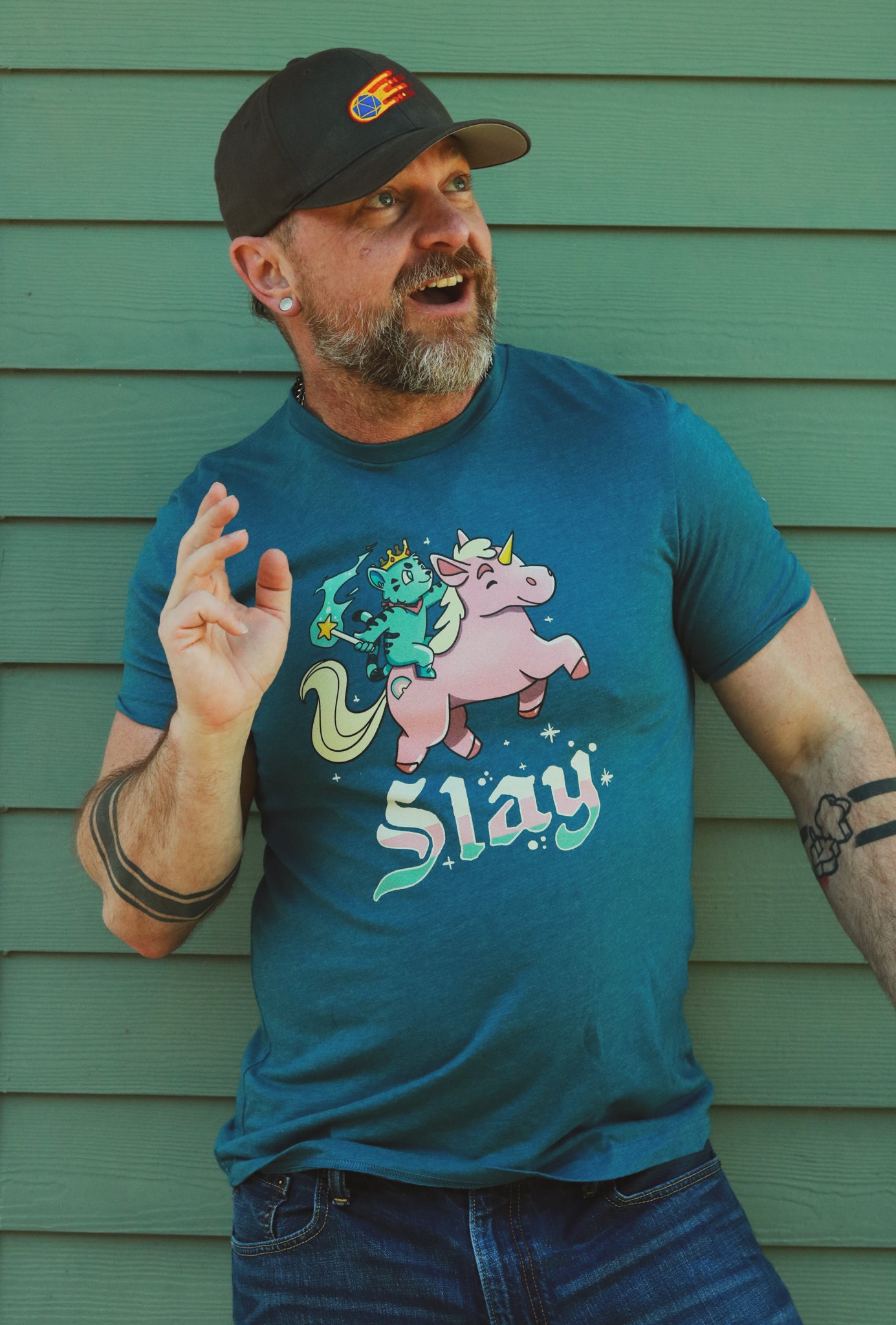 a light blue tank top with blue tiger riding pink unicorn, with the words slay below the image, worn by a surprised bearded hairy guy.